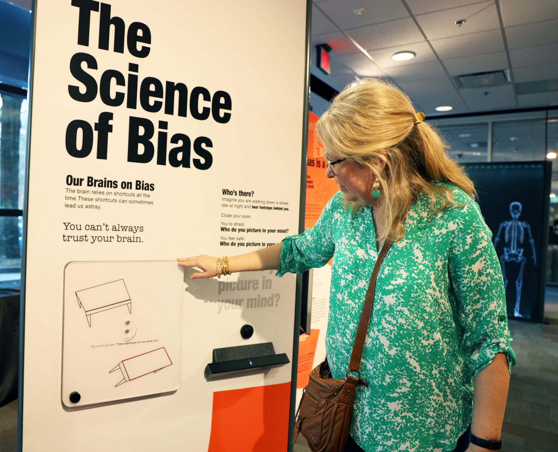 A woman in a green blouse points at a museum display titled "The Science of Bias," which discusses how our brains can be tricked. The display features text and an illustration of two tables.