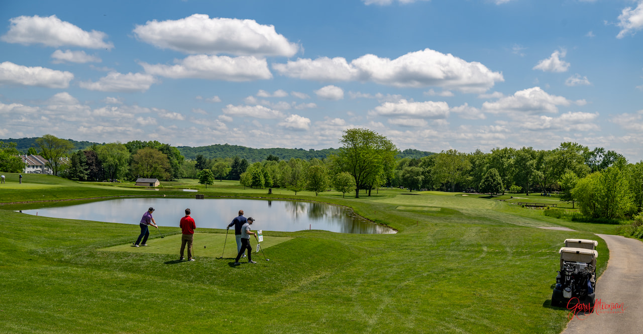 Three people playing golf on a lush green course with a small pond nearby under a partly cloudy sky. A golf cart is parked on the right. Trees and hills are visible in the distance.