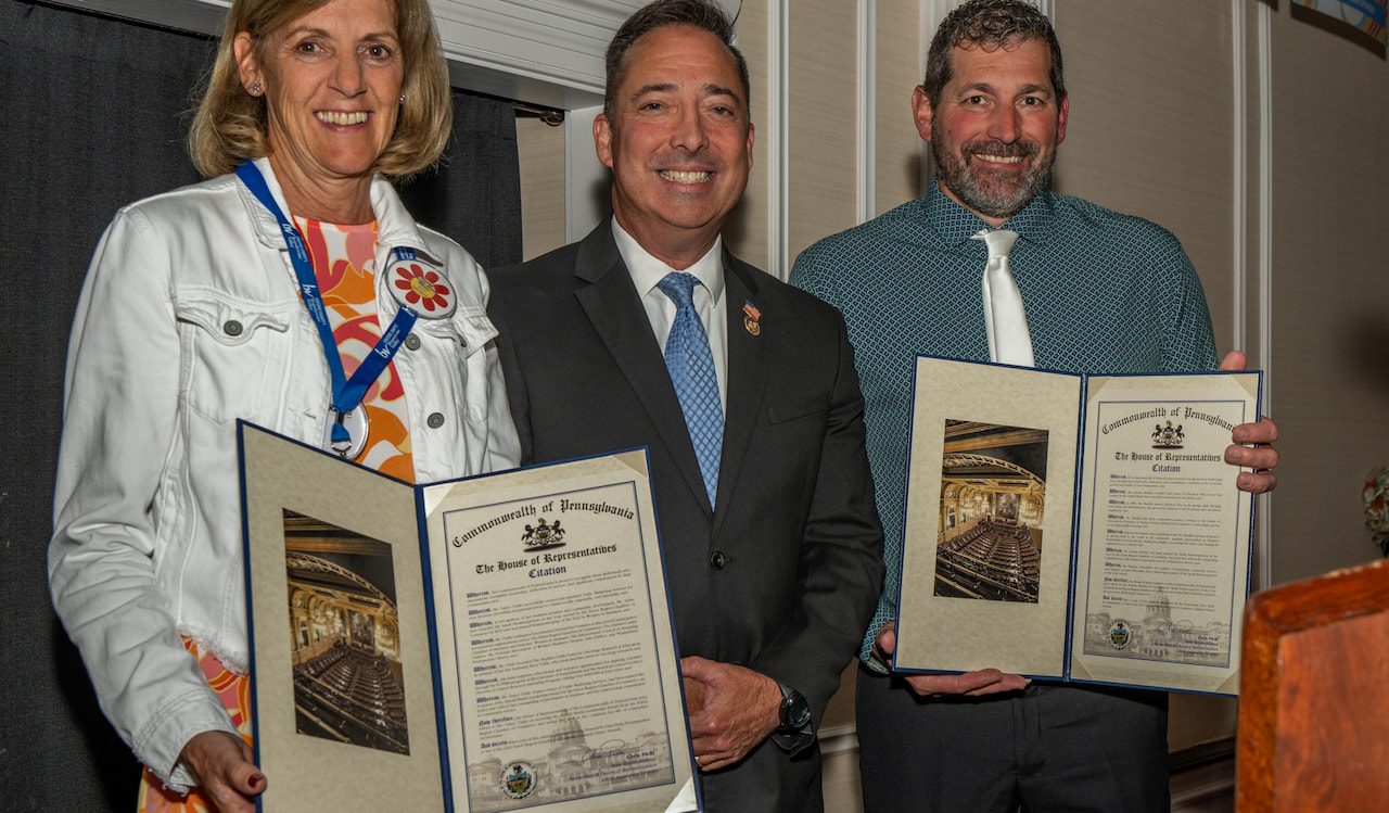 Three people smiling and holding framed certificates at an event. The certificates have official-looking seals at the bottom. The person on the left is wearing a white jacket, the middle person a dark suit, and the person on the right a teal shirt with a tie.