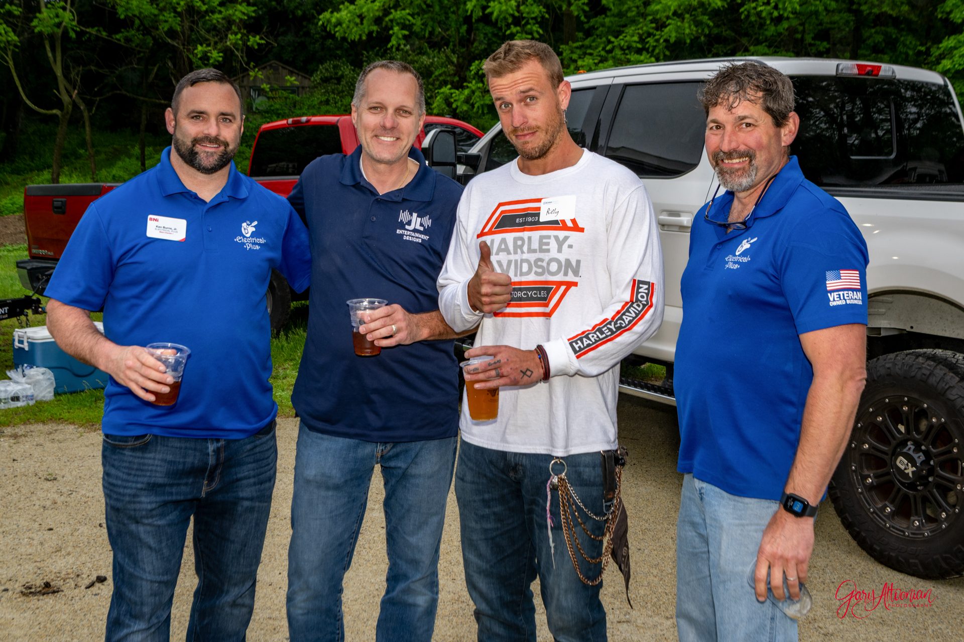 Four men stand outdoors in front of vehicles, smiling at the camera. Three wear blue shirts and name tags, while one in the middle wears a white Harley-Davidson shirt. They are holding drinks and appear to be at a casual gathering.