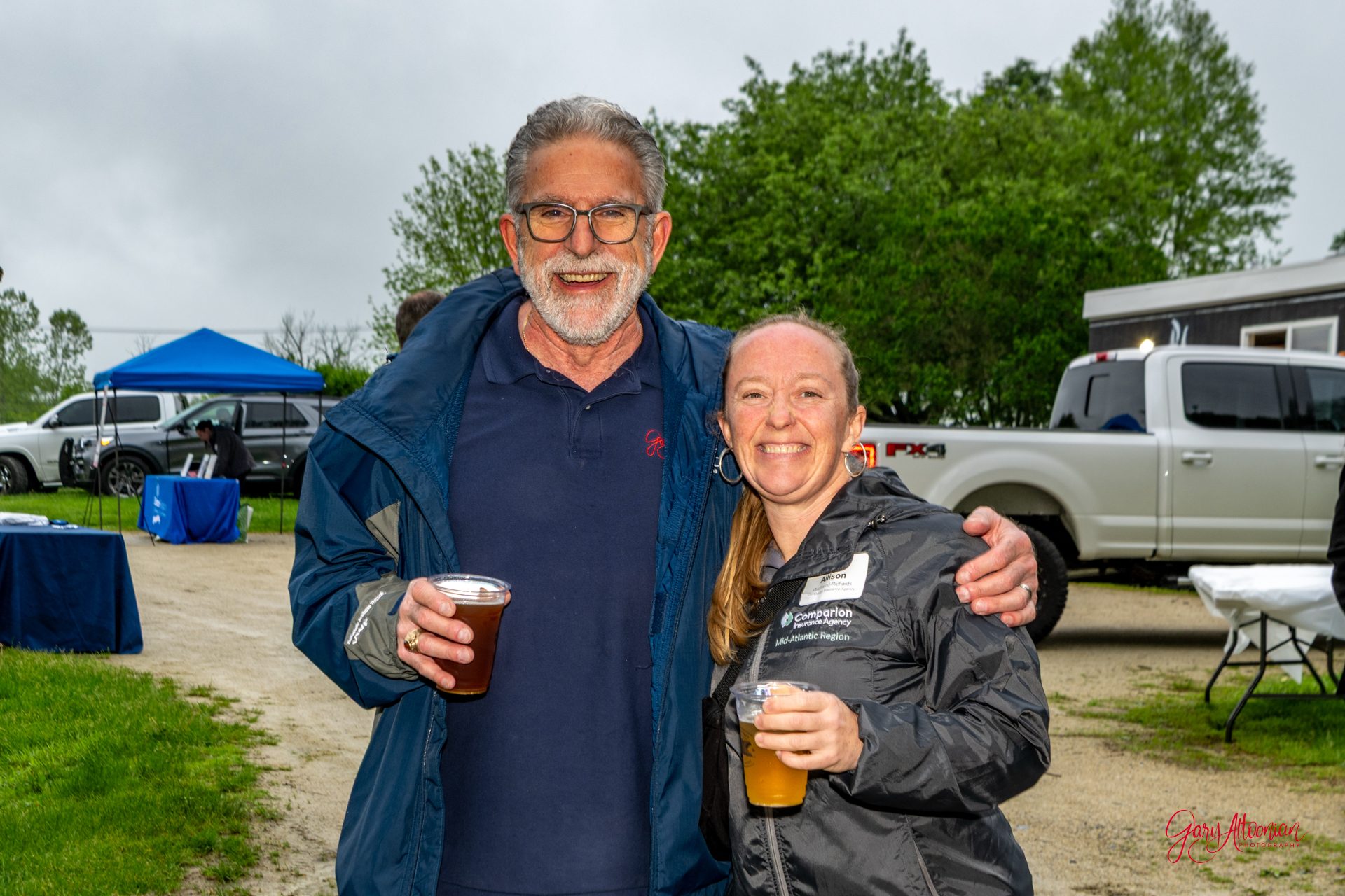 A smiling man and woman stand outdoors with drinks in hand, wearing jackets. The man has gray hair and glasses, and the woman has a name tag. Vehicles and tents are visible in the background on a cloudy day.
