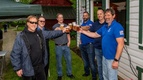 Six men standing outside near buildings, smiling and raising glasses of beer in a toast; most are wearing casual clothes and two are in blue shirts with logos. The setting appears to be a casual outdoor gathering.