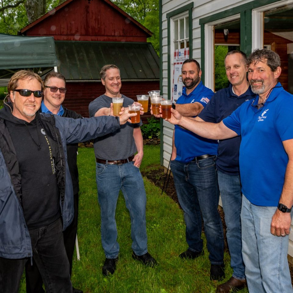 Six men standing outside near buildings, smiling and raising glasses of beer in a toast; most are wearing casual clothes and two are in blue shirts with logos. The setting appears to be a casual outdoor gathering.
