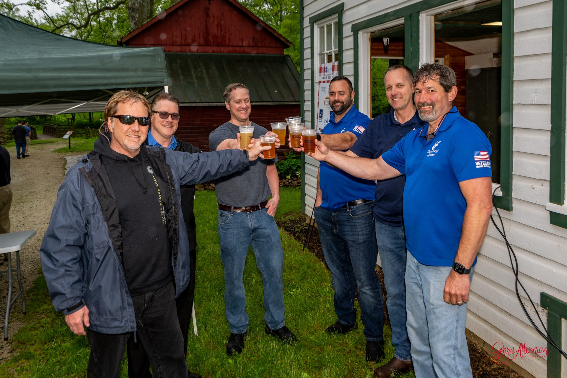 Six men standing outside near buildings, smiling and raising glasses of beer in a toast; most are wearing casual clothes and two are in blue shirts with logos. The setting appears to be a casual outdoor gathering.