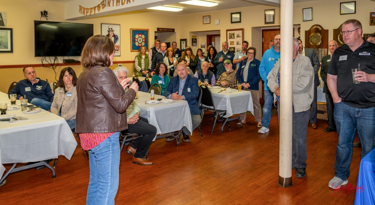 A woman stands and speaks to a group of people seated and standing in a decorated room with tables, listening attentively. There are banners, framed items, and a large TV on the walls.