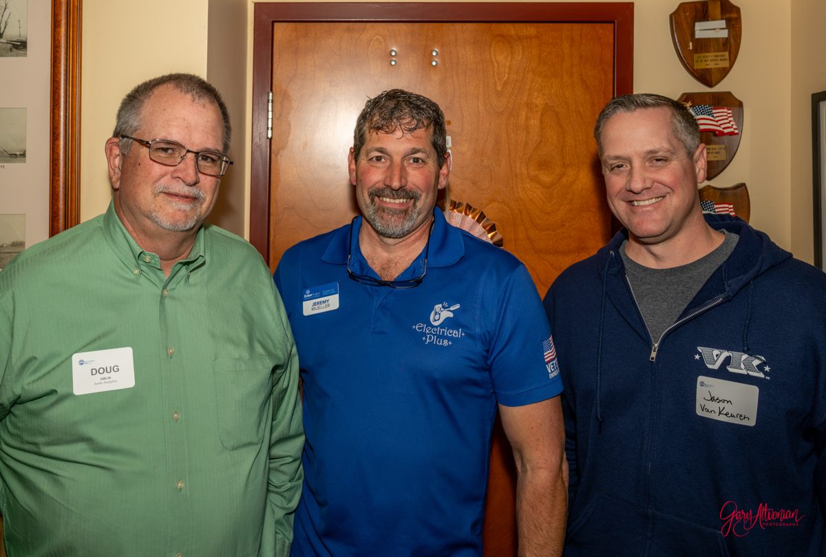 Three men standing indoors, smiling for a photo. The man on the left wears glasses and a green shirt, the middle man wears a blue polo shirt, and the man on the right wears a dark hoodie. Plaques are visible on the wall behind them.
