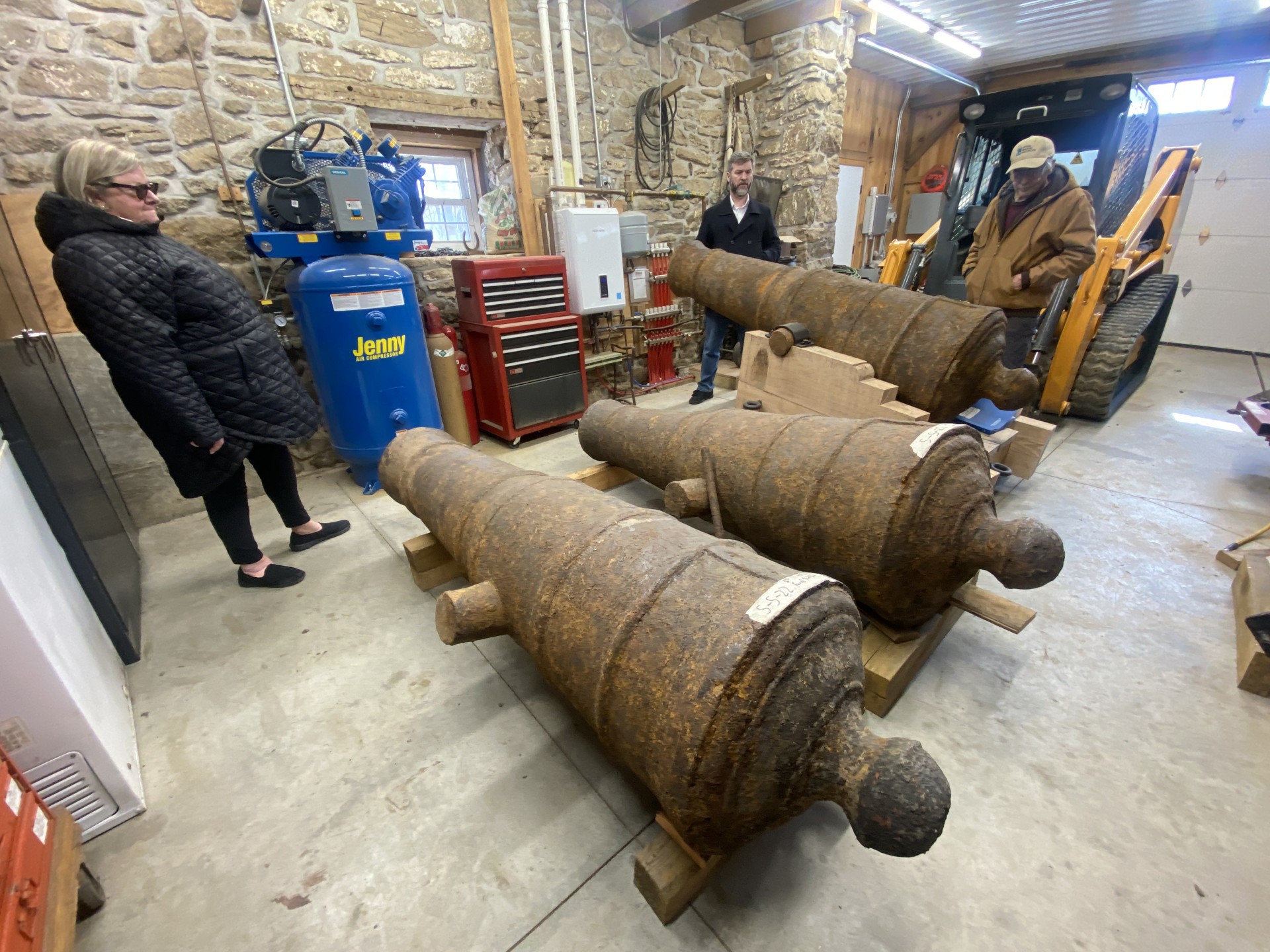 Three rusty cannons are displayed in a garage. Two people stand nearby observing them, while another person is on a mini loader. A stone wall and various tools surround the scene.