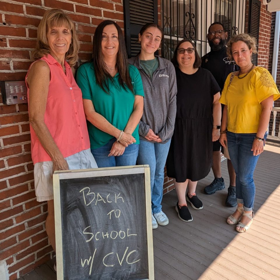 A group of six people stand on a porch in front of a brick building. They are smiling and standing behind a chalkboard sign that reads, "BACK TO SCHOOL w/ CVC." The group includes four women and two men, all dressed in casual attire.