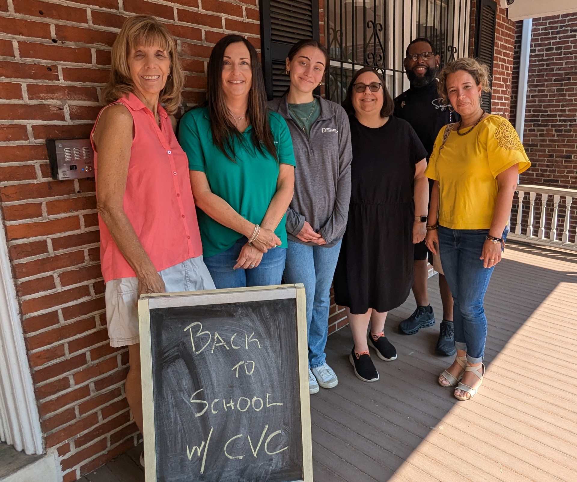 A group of six people stand on a porch in front of a brick building. They are smiling and standing behind a chalkboard sign that reads, "BACK TO SCHOOL w/ CVC." The group includes four women and two men, all dressed in casual attire.