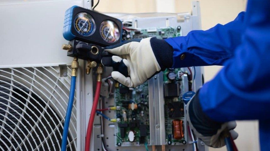 A technician in blue coveralls and gloves uses a digital manifold gauge to check the refrigerant levels on an outdoor air conditioning unit with exposed circuit boards.