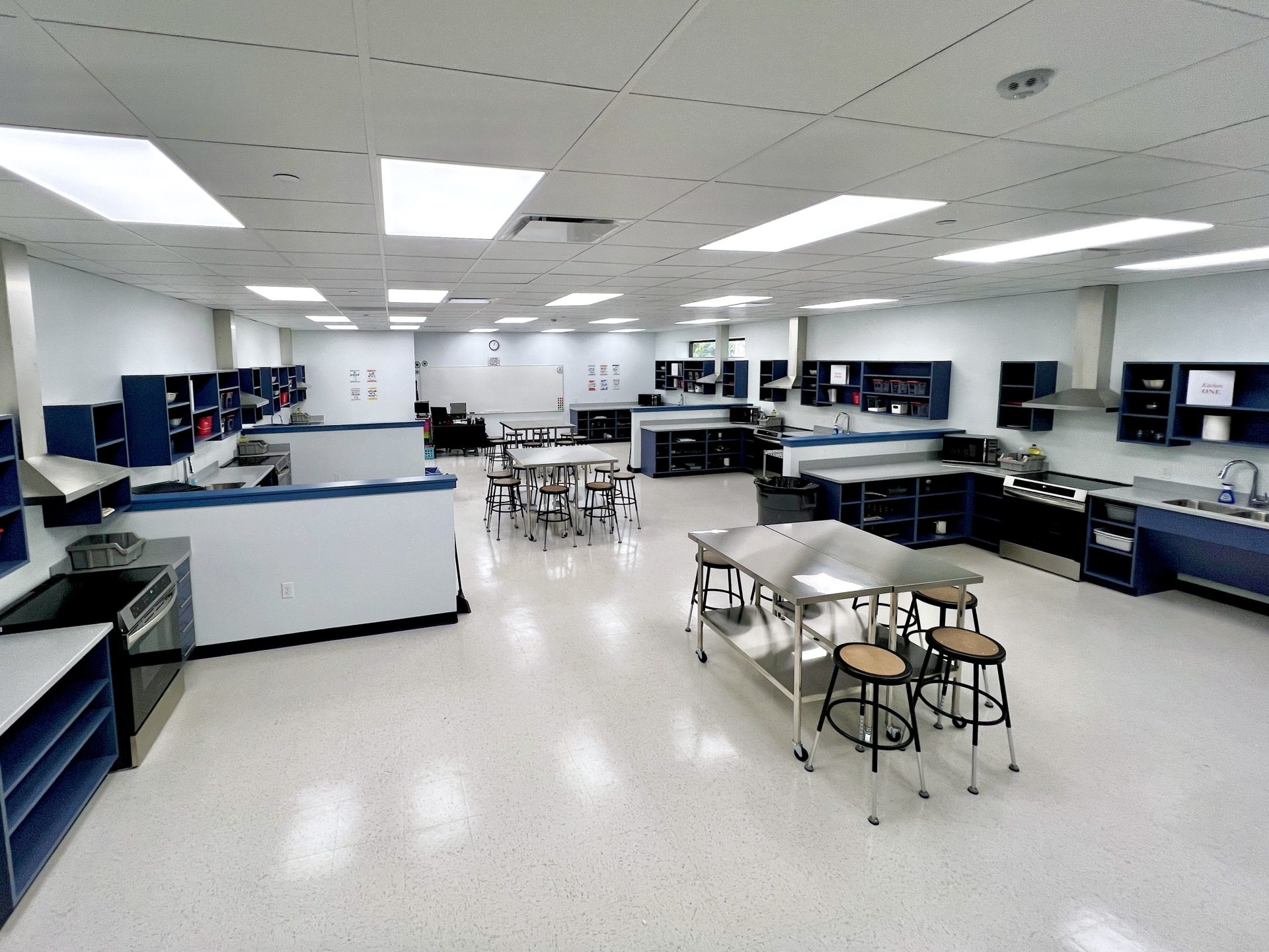 A spacious, modern classroom kitchen with multiple cooking stations, stainless steel tables, blue shelving, ovens, and sinks. Several stools and tables are arranged in the center, and posters are on the far wall.
