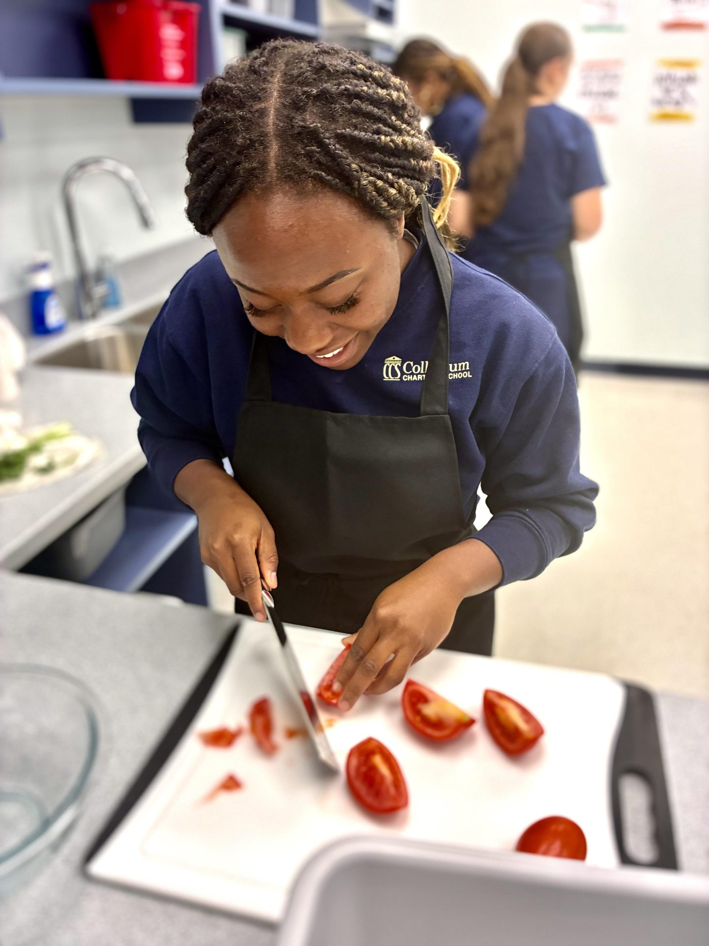 A woman wearing a dark apron smiles while slicing tomatoes on a white cutting board in a kitchen. Two other people are blurred in the background, and kitchen counters and sinks are visible.