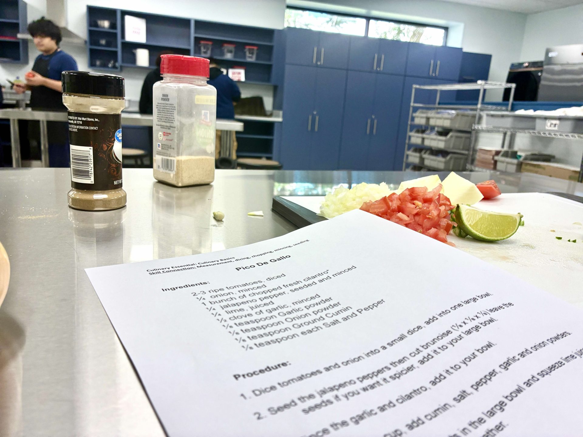 A kitchen with people cooking in the background, a stainless steel counter with spice jars, chopped vegetables, a lime, and a printed Pico de Gallo recipe in the foreground.