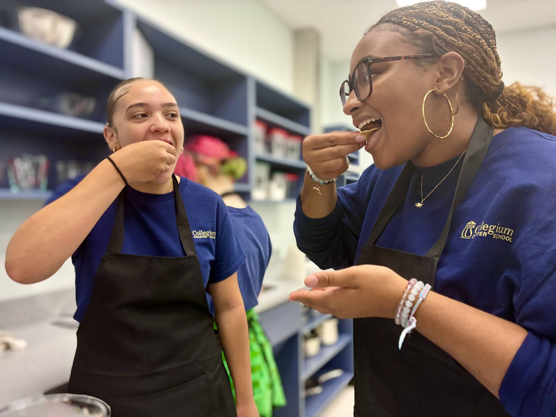Two young women wearing aprons and blue shirts stand in a kitchen, smiling as they taste food from their hands. Shelves with bowls and kitchen supplies are visible in the background.