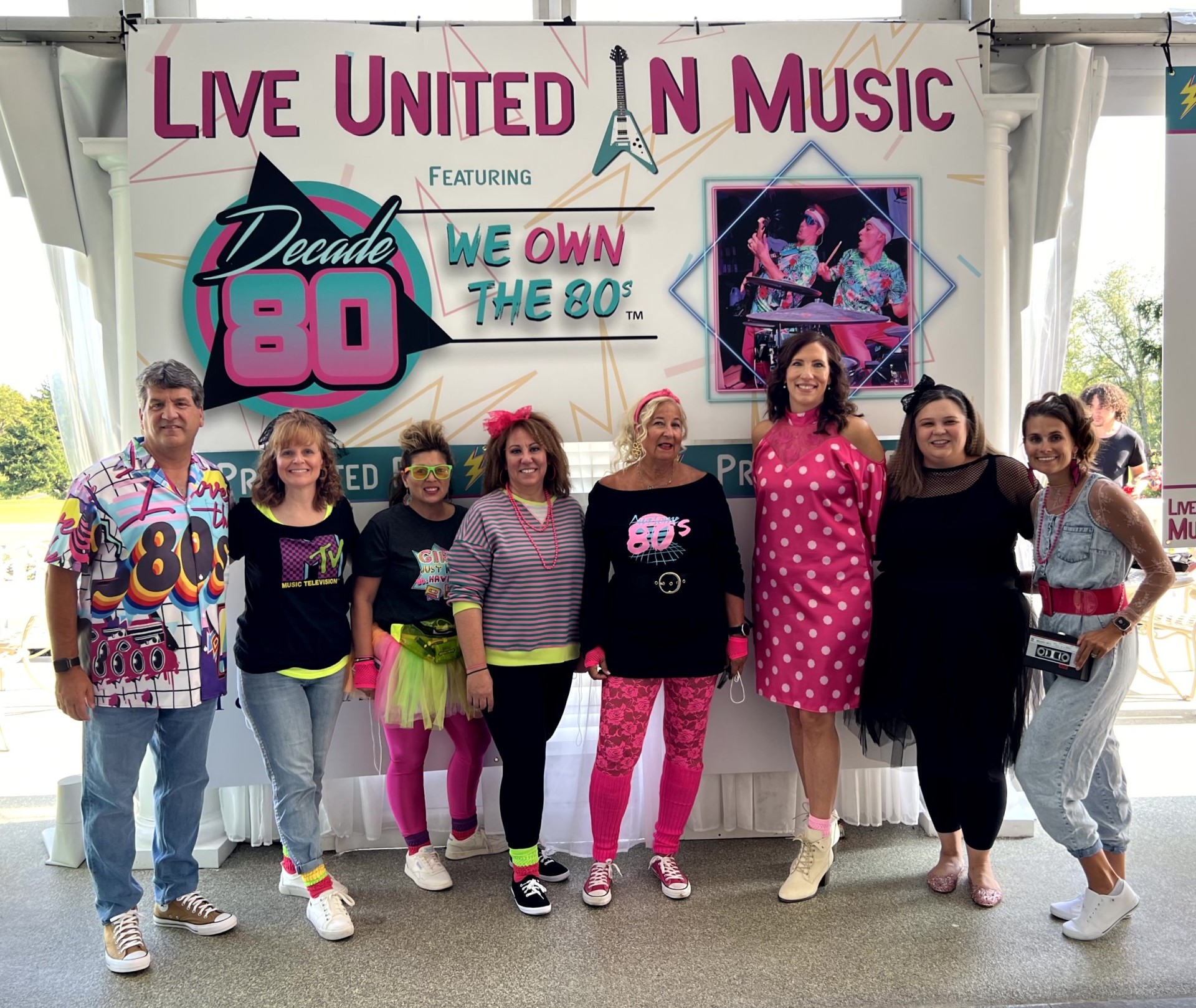 A group of people dressed in 80s-themed clothing stand together in front of a "Live United in Music" banner with neon designs. They are smiling and appear to be at an event celebrating 1980s music.