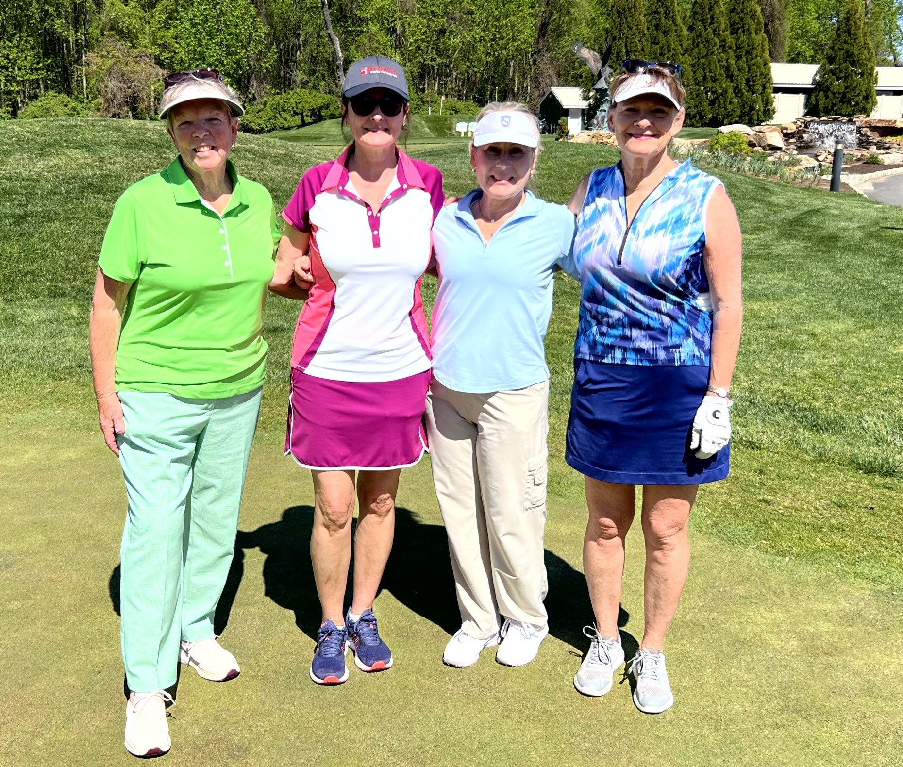 Four women in golf attire stand together on a green golf course, smiling at the camera on a sunny day. Trees and a building are visible in the background.