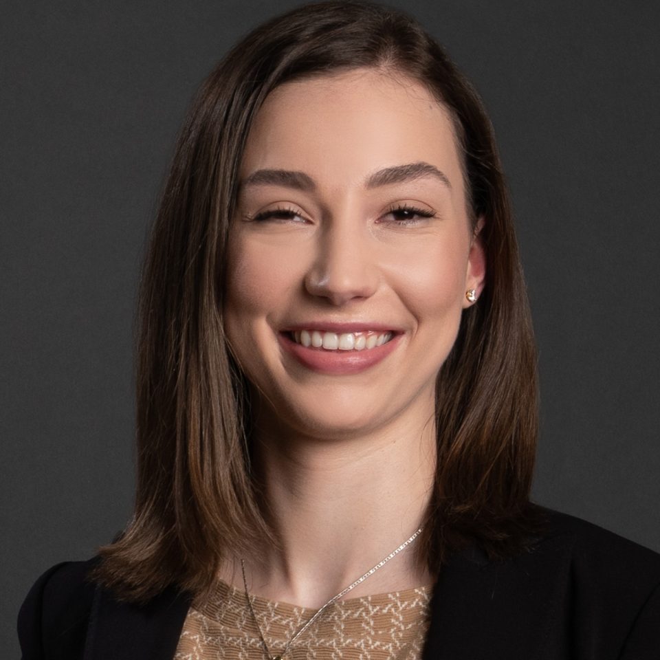 A professional headshot of a smiling woman with shoulder-length brown hair, wearing a black blazer and a lace top.