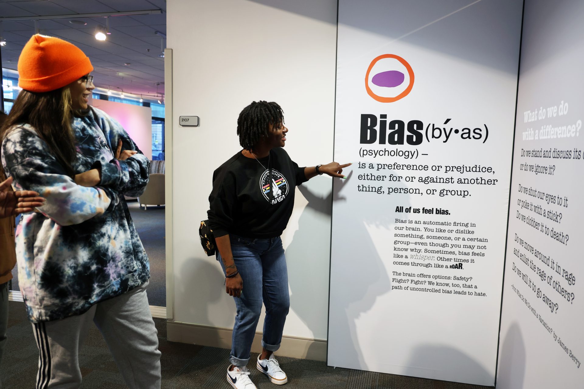 Two women stand in front of an exhibit panel titled "Bias," which defines the term and explains that everyone feels bias. One woman points at the text while both appear engaged and interested in the display.