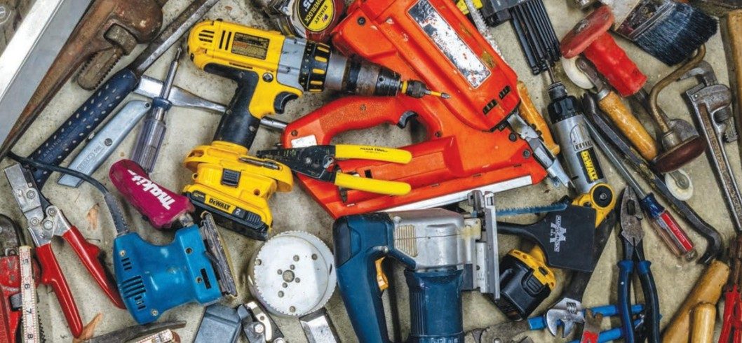 A variety of hand and power tools, including drills, a saw, pliers, hammers, utility knives, and screwdrivers, are scattered on a work surface in a messy arrangement.