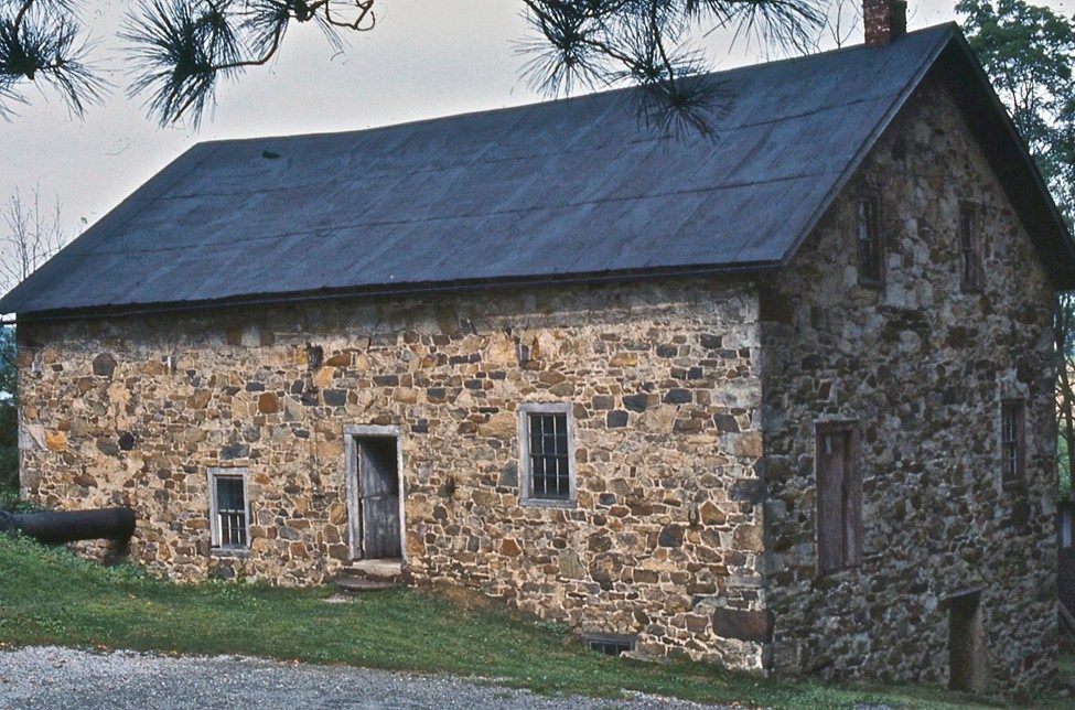 A rustic stone building with a dark, sloped roof, wooden door, and small windows sits on a grassy slope. Trees and branches are visible in the background and foreground.